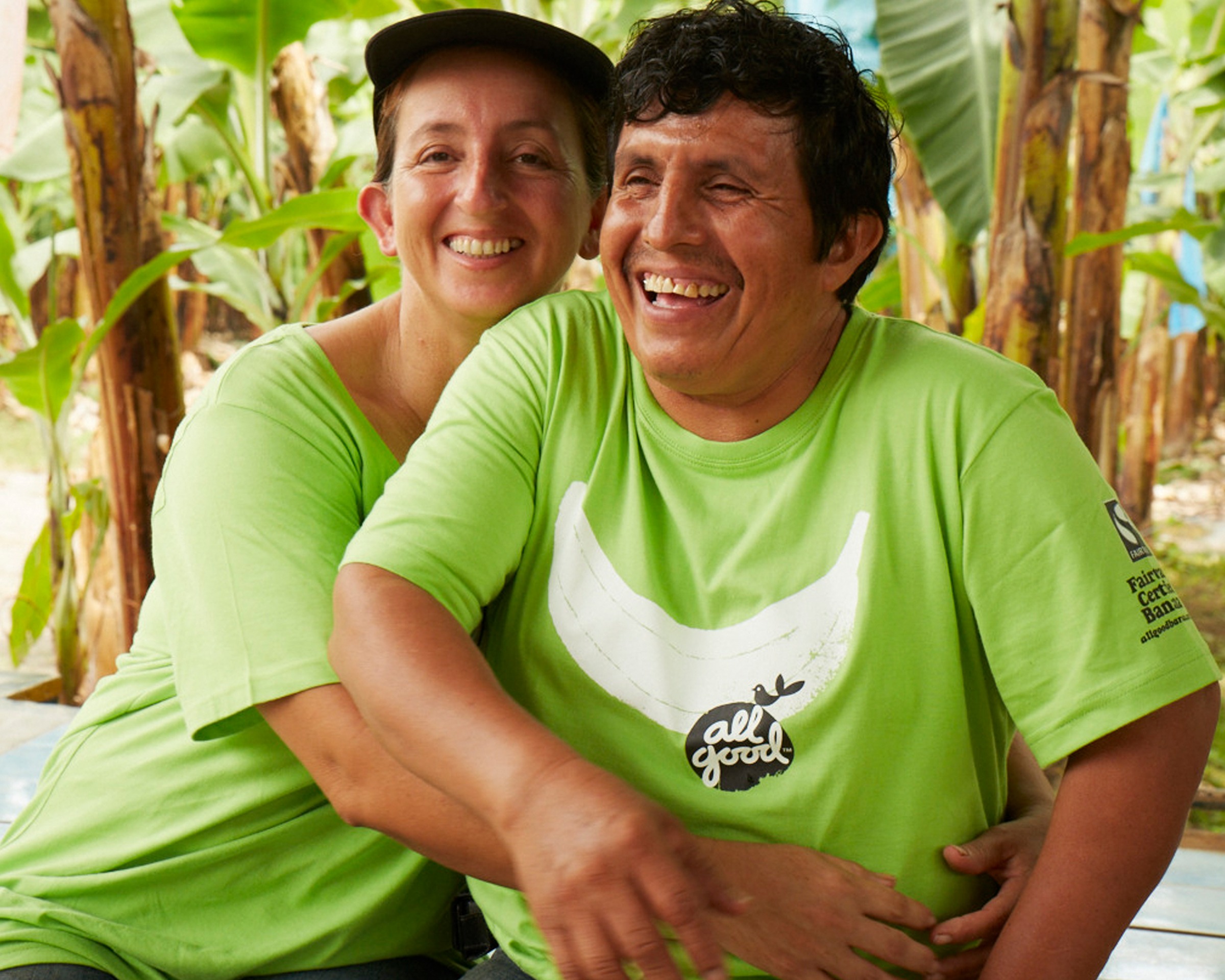 Two people in green All Good t-shirts smiling warmly, one with their arm around the other, banana plants in the background