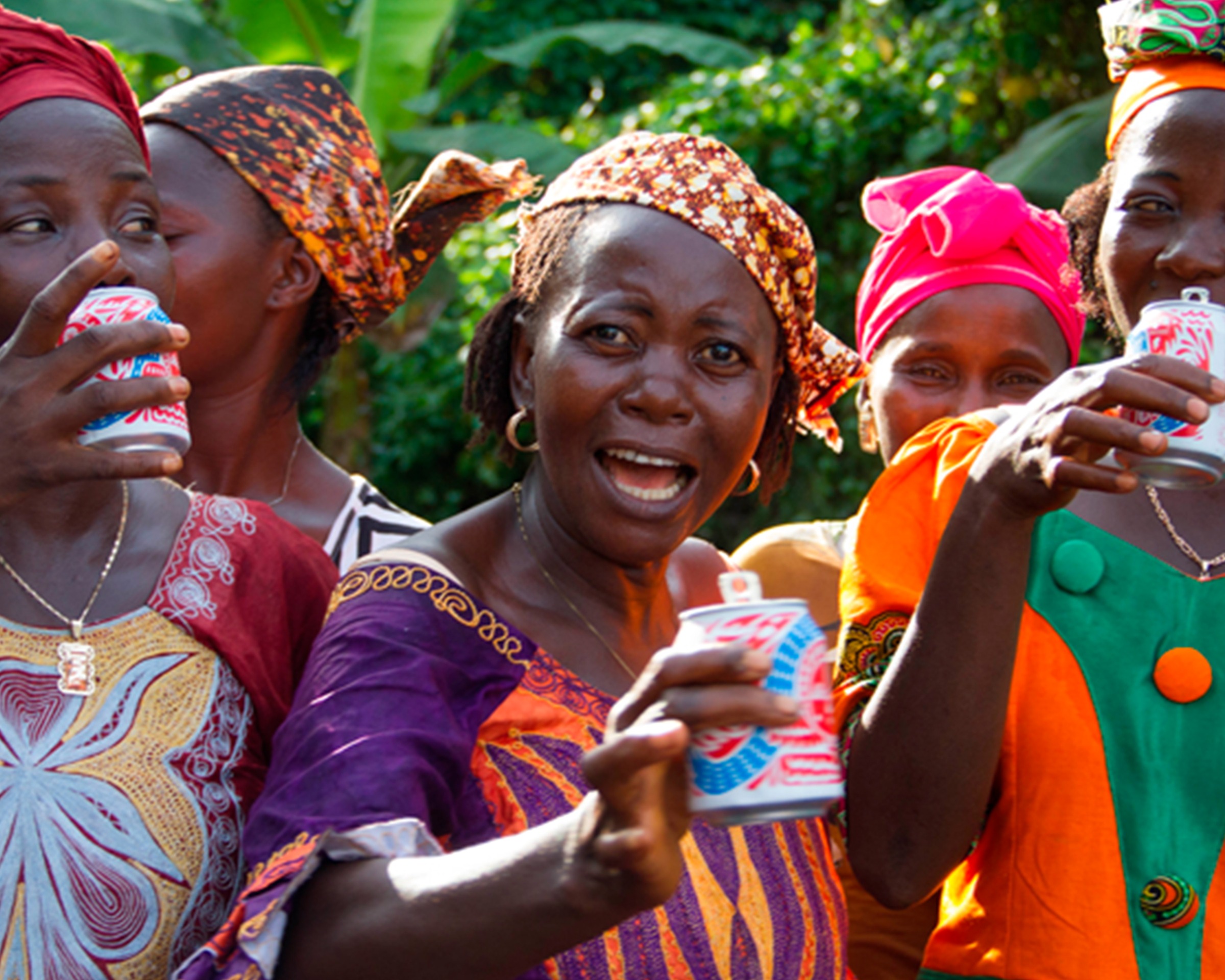 Sierra Leone women in headscarves and patterned dress drinking Karma Cola, in front of banana leaves