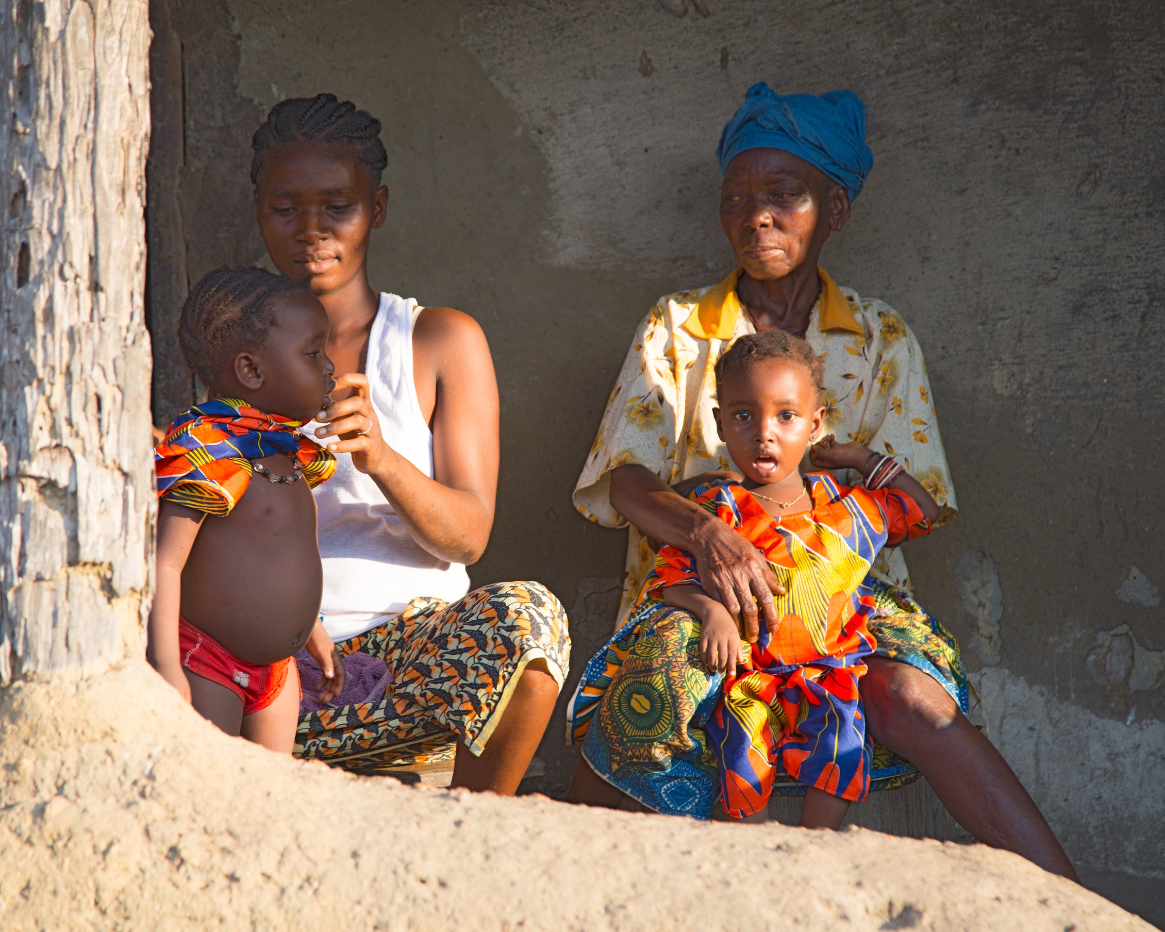 A mother feeding her young child and an older woman holding another small child, sitting on the threshold of a mud-walled home in Sierra Leone