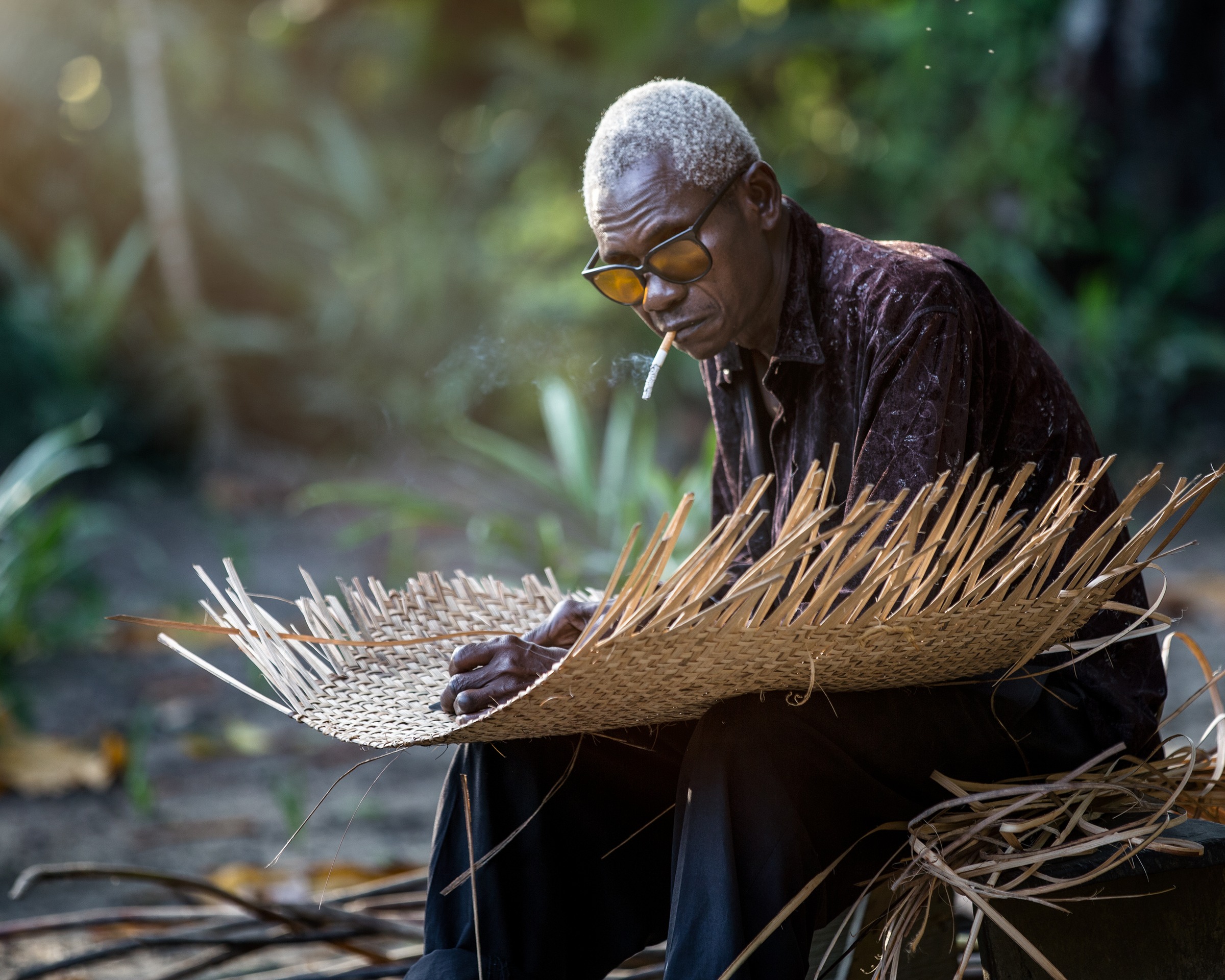 An older man in dark glasses, cigarette in mouth, weaving a wide palm-frond mat in golden afternoon light