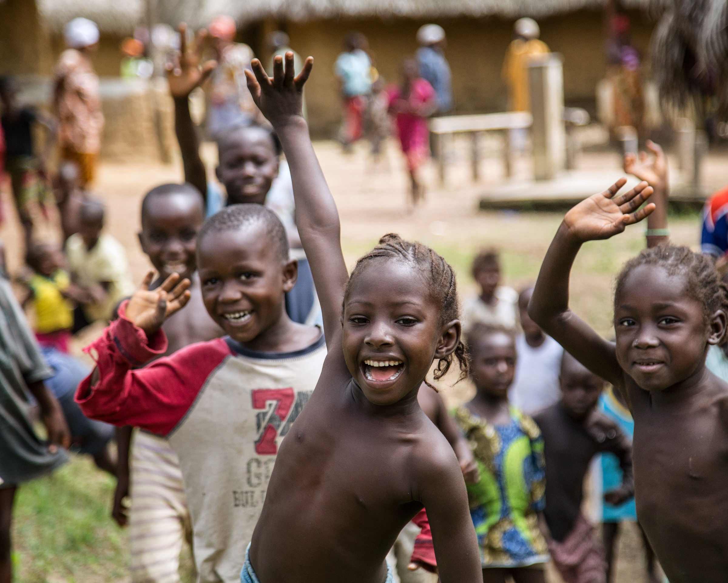 A group of Sierra Leonean children in a village, hands raised mid-cheer, smiling broadly