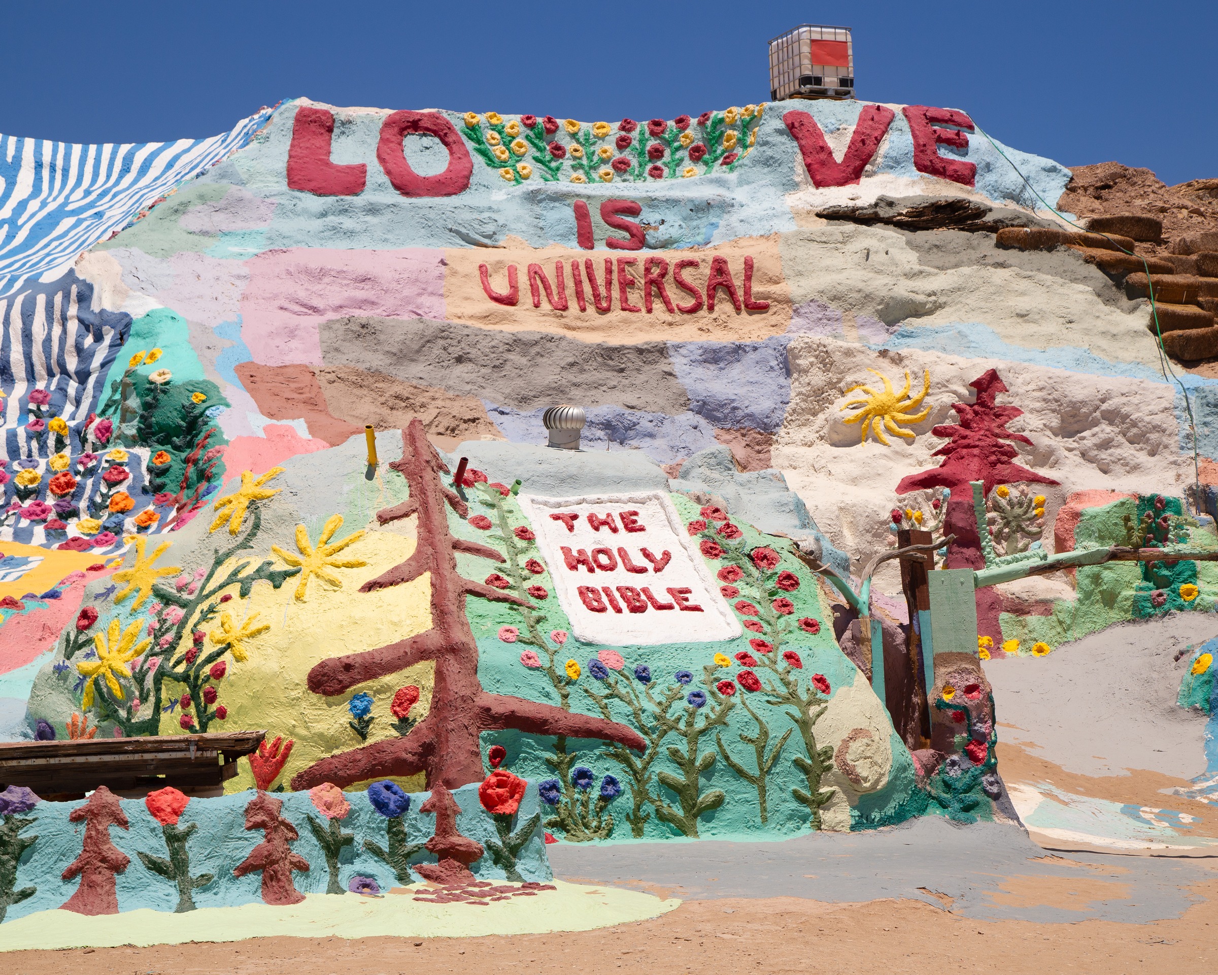 Painted detail of the mountain — LOVE IS UNIVERSAL above a Holy Bible motif surrounded by hand-formed flowers and trees