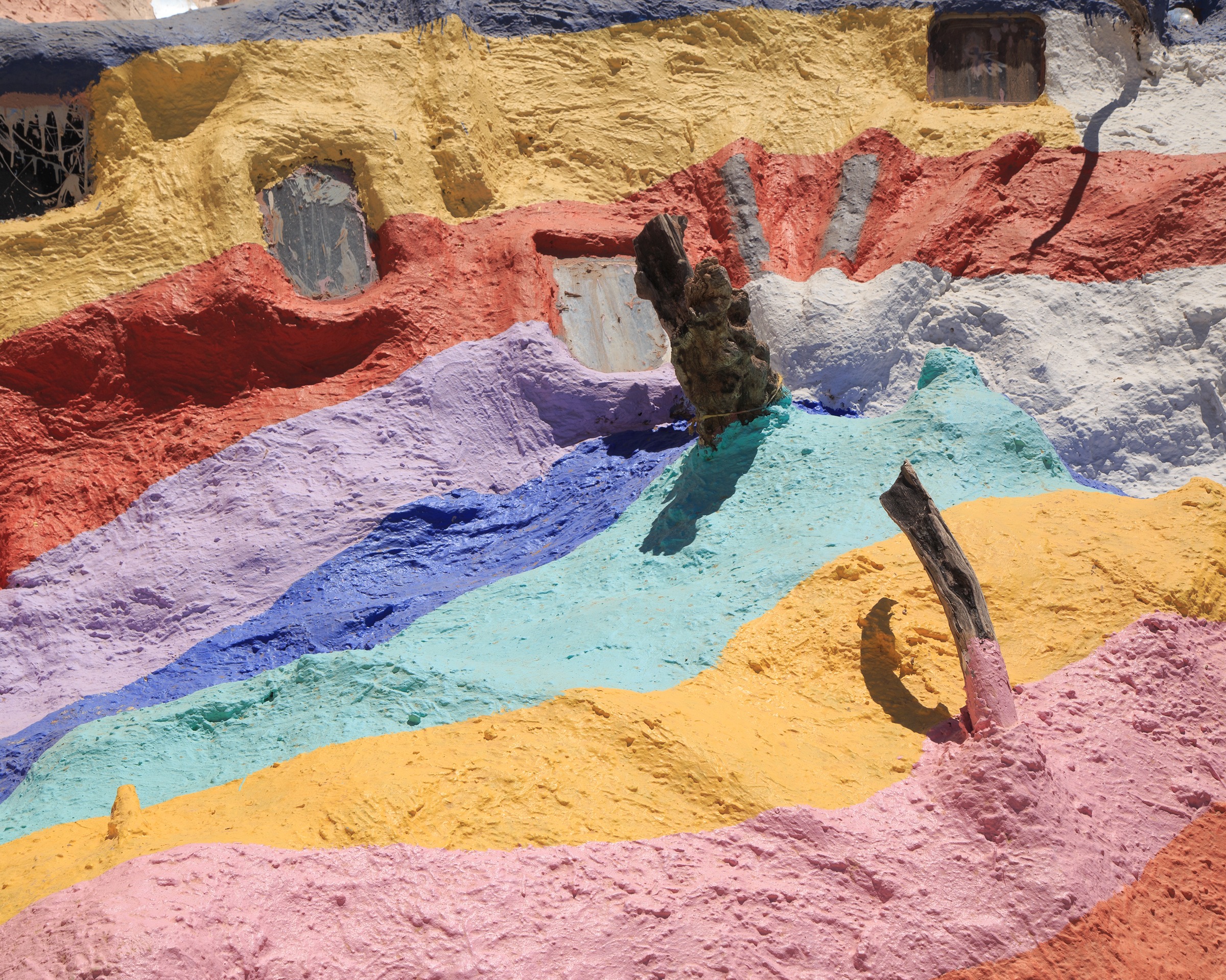Pure colour study — bands of yellow, red, lavender, blue and orange painted across the mountain face with desert detritus catching the light